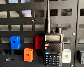 May include: A black two-way radio with an antenna, mounted on a black metal grid wall. The radio is attached to a white plastic hook. There are three small plastic hooks, one blue, one orange, and one red, also mounted on the wall.