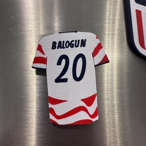 Puede incluir: Un im&aacute;n blanco con forma de camiseta con detalles rojos y azules. La camiseta presenta el nombre "BALOGUN" sobre el n&uacute;mero "20" en azul marino. Rayas rojas adornan las mangas y la parte inferior.
