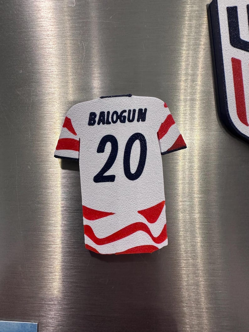 Puede incluir: Un im&aacute;n blanco con forma de camiseta con detalles rojos y azules. La camiseta presenta el nombre "BALOGUN" sobre el n&uacute;mero "20" en azul marino. Rayas rojas adornan las mangas y la parte inferior.