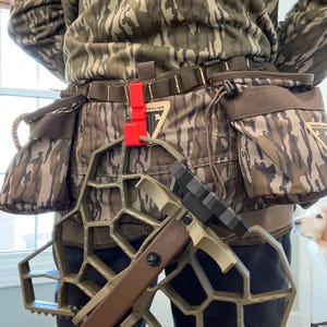 May include: A camouflage hunting belt with attached pouches and a climbing system. The climbing system is olive green and brown, with a black footrest. A red clip is attached to the belt. The person is wearing a camouflage hoodie.