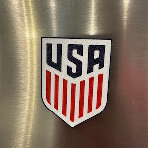 Puede incluir: Un parche en forma de escudo blanco, rojo y azul con las letras "USA" en azul marino en la parte superior y rayas verticales rojas debajo. Es probable que sea un emblema de un equipo deportivo o un art&iacute;culo patri&oacute;tico.