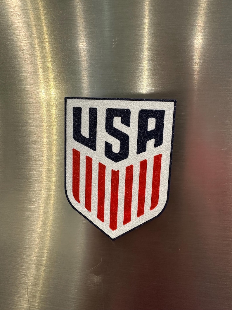 Puede incluir: Un parche en forma de escudo blanco, rojo y azul con las letras "USA" en azul marino en la parte superior y rayas verticales rojas debajo. Es probable que sea un emblema de un equipo deportivo o un art&iacute;culo patri&oacute;tico.