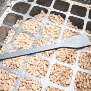 May include: A black plastic tool for moving wood pellets in a pellet grill. The tool is lying on top of a metal grate filled with wood pellets.