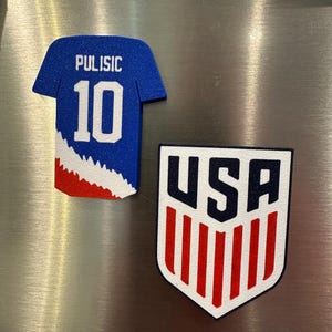 Puede incluir: Dos imanes para nevera. Uno tiene forma de camiseta de f&uacute;tbol azul con "Pulisic 10" en blanco. El otro es un escudo con "USA" en negro y rayas rojas y blancas.