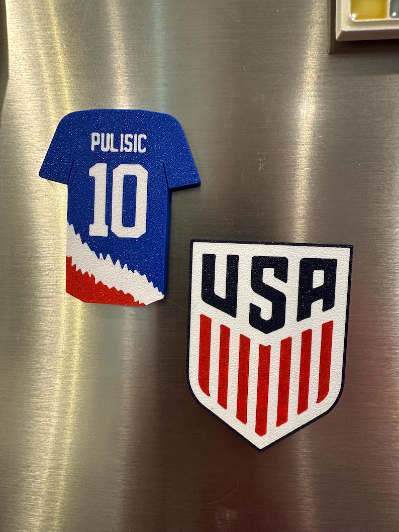 Puede incluir: Dos imanes para nevera. Uno tiene forma de camiseta de f&uacute;tbol azul con "Pulisic 10" en blanco. El otro es un escudo con "USA" en negro y rayas rojas y blancas.