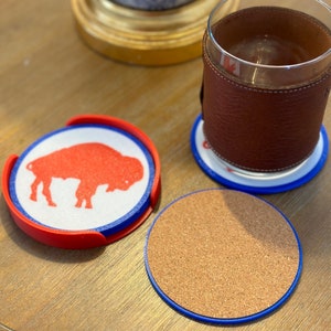 May include: Three round coasters, one with a red buffalo silhouette on a white background, one with a cork surface, and one with a blue and white design. All coasters have a blue outer ring.