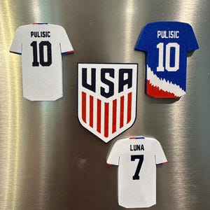 Puede incluir: Un conjunto de imanes con tem&aacute;tica de f&uacute;tbol, con el logotipo de USA y camisetas. Las camisetas son blancas y azules, con los nombres "Pulisic" y "Luna" y los n&uacute;meros 10 y 7. El logotipo de USA es rojo, blanco y azul.