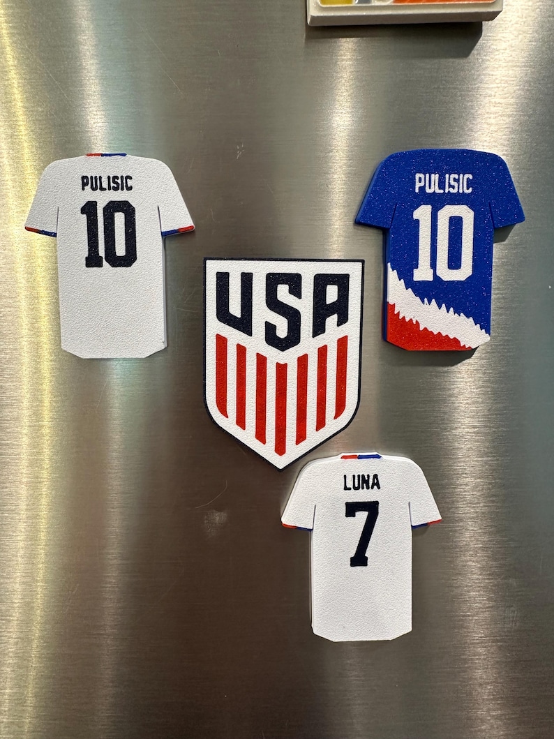 Puede incluir: Un conjunto de imanes con tem&aacute;tica de f&uacute;tbol, con el logotipo de USA y camisetas. Las camisetas son blancas y azules, con los nombres "Pulisic" y "Luna" y los n&uacute;meros 10 y 7. El logotipo de USA es rojo, blanco y azul.