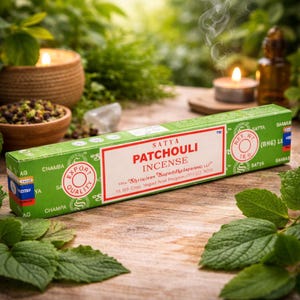 Peut inclure: Une boîte verte d'encens Satya Patchouli avec une étiquette rouge et blanche. La boîte est posée sur une surface en bois avec des feuilles vertes et des bougies allumées en arrière-plan. De la fumée s'élève de l'encens.