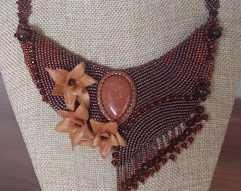 May include: A handcrafted necklace featuring a central oval-shaped, sparkling, brown stone. The necklace is made of small, brown beads, with a floral accent and a dangling teardrop-shaped bead. The necklace is displayed on a mannequin.