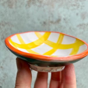 以下が含まれることがあります： 手描きの小さなセラミック皿。内側は白で黄色の格子模様、縁はオレンジ色、外側は緑色です。皿は手で持たれており、そのサイズとデザインを示しています。
