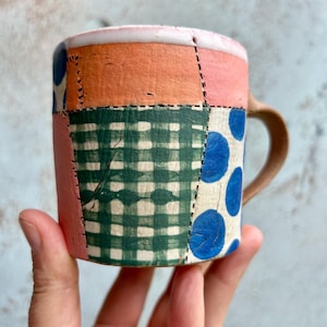 Pode incluir: Caneca de cerâmica artesanal com um design de patchwork. A caneca apresenta secções de laranja, rosa, xadrez verde e branco com bolinhas azuis. A pega é de cor castanho claro. A caneca é segurada por uma mão.
