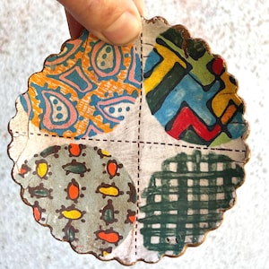 Puede incluir: Un posavasos redondo hecho a mano con un borde festoneado, dividido en cuatro secciones. Cada sección presenta un patrón colorido único: cachemira, formas abstractas, pequeñas figuras parecidas a tortugas y un diseño de cuadros verdes. El posavasos está sostenido por una mano.
