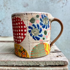 Puede incluir: Una taza de cerámica hecha a mano con un asa marrón y un diseño de patchwork. La taza presenta lunares rojos, patrones florales azules y formas geométricas amarillas y verdes. La taza está sobre una superficie de madera.