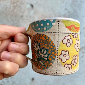 Pode incluir: Caneca de cerâmica artesanal com um design de patchwork único. A caneca apresenta padrões coloridos, incluindo bolinhas azuis, círculos laranja e flores amarelas. A alça é de cor marrom natural e a caneca é segurada por uma mão.