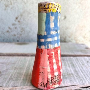 May include: A handcrafted ceramic vase with a distinctive, vibrant design. The vase showcases a red base with white vertical stripes, a blue and white chequered middle section, and a yellow and black patterned top. The initials "SM" are visible.