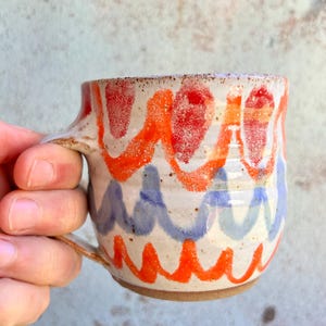 May include: Handmade ceramic mug with a cream base and a wavy pattern in red, orange, and blue. The mug has a brown handle and base. A unique piece for coffee or tea.