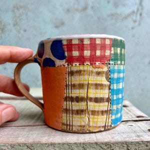 Pode incluir: Uma caneca de cerâmica patchwork com uma alça castanha. O exterior da caneca apresenta uma variedade de padrões de tecido, incluindo bolinhas azuis, xadrez vermelho e xadrez amarelo. A caneca tem aproximadamente 7.6 cm de altura.