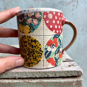Pode incluir: Uma pequena caneca de cerâmica com um design de patchwork. A caneca tem uma alça castanha e é decorada com padrões florais e de bolinhas coloridas. A caneca tem cerca de 7,6 cm de altura.