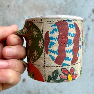 Pode incluir: Uma pequena caneca feita à mão com um design de patchwork único. A caneca apresenta uma variedade de padrões coloridos pintados à mão, incluindo formas vermelhas, azuis e verdes. A alça está integrada no design. A caneca tem aproximadamente 7.6 cm de altura.