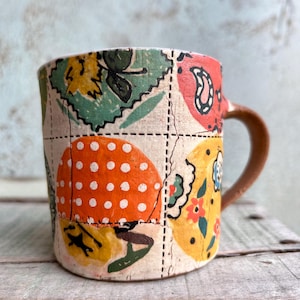 Pode incluir: Uma caneca de cerâmica com um design de patchwork com ilustrações coloridas de cogumelos, flores e folhas. A caneca tem uma alça marrom e está colocada sobre uma superfície de madeira rústica.