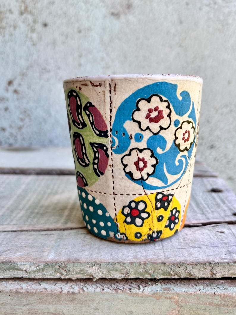 May include: A small, handmade cup with a patchwork design. The cup has a cream base with sections of blue, yellow, and green, each decorated with floral and paisley patterns. The cup is set on a weathered wooden surface.