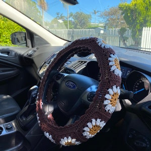 May include: A brown and white crocheted steering wheel cover with a daisy pattern. The cover is on a black steering wheel with the word "Ford" in white lettering.