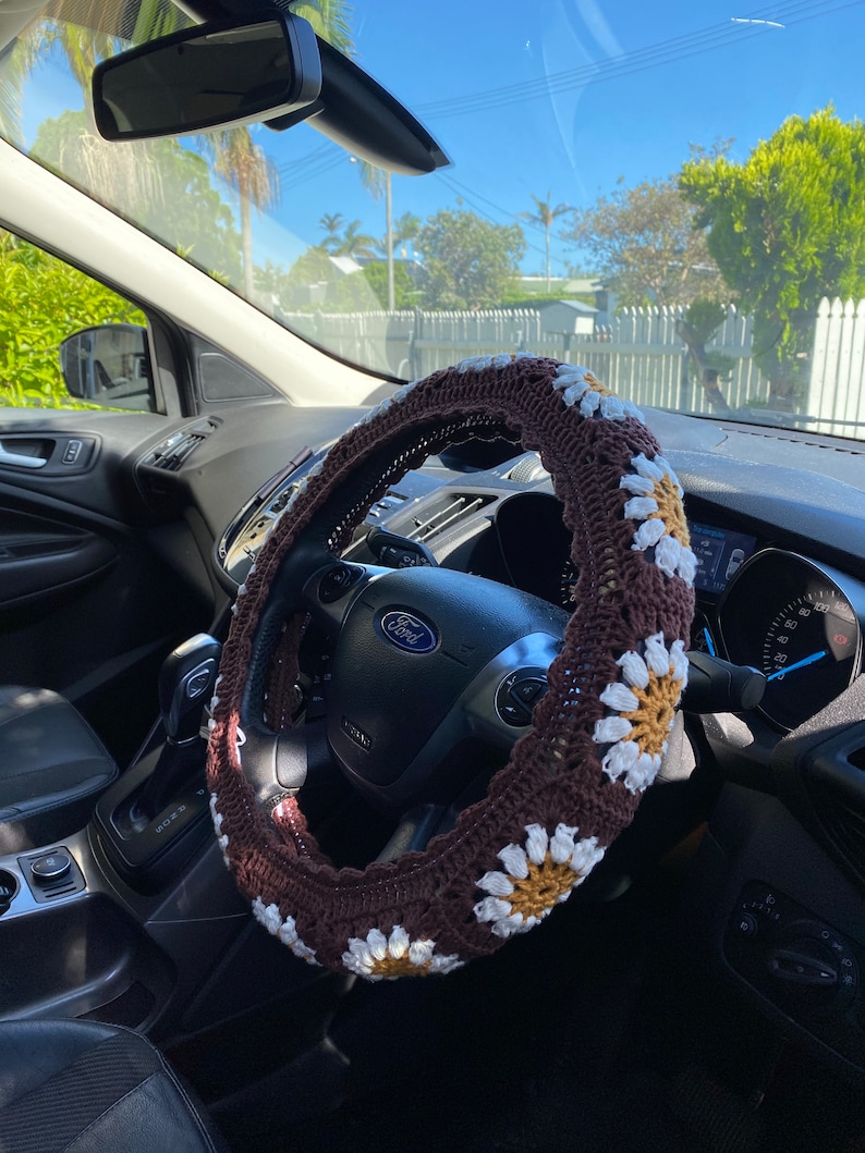 May include: A brown and white crocheted steering wheel cover with a daisy pattern. The cover is on a black steering wheel with the word "Ford" in white lettering.