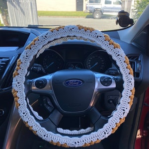 May include: A white and gold crochet steering wheel cover. The cover is crocheted with a delicate pattern and has a white base with gold accents. The cover is designed to fit over a standard steering wheel.