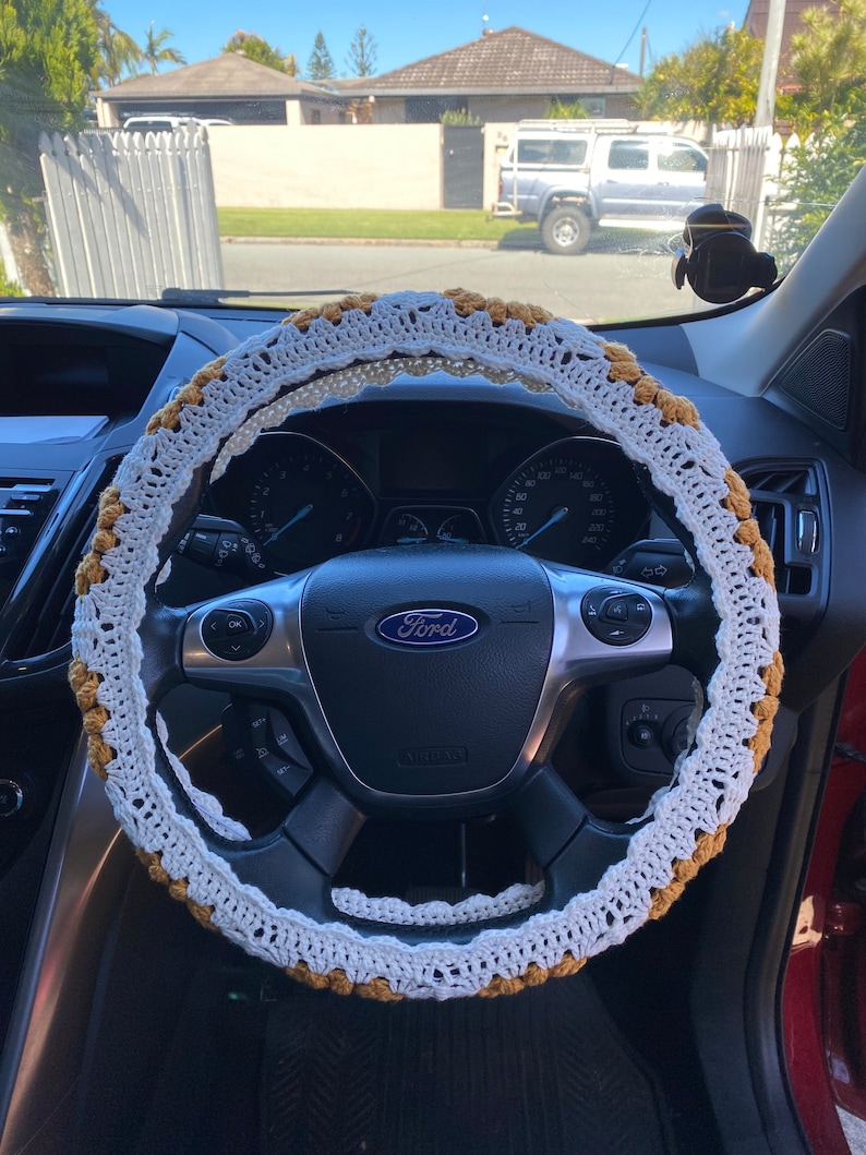 May include: A white and gold crochet steering wheel cover. The cover is crocheted with a delicate pattern and has a white base with gold accents. The cover is designed to fit over a standard steering wheel.