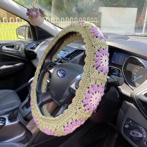 May include: A crocheted steering wheel cover in green and purple with a flower pattern. The cover is on a car steering wheel with the Ford logo visible.