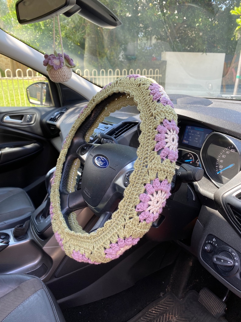 May include: A crocheted steering wheel cover in green and purple with a flower pattern. The cover is on a car steering wheel with the Ford logo visible.