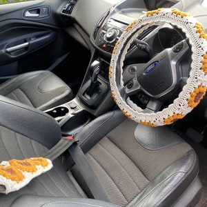 May include: The interior of a black car with a crocheted steering wheel cover. The cover is white with brown accents. The car has black leather seats and a black dashboard.