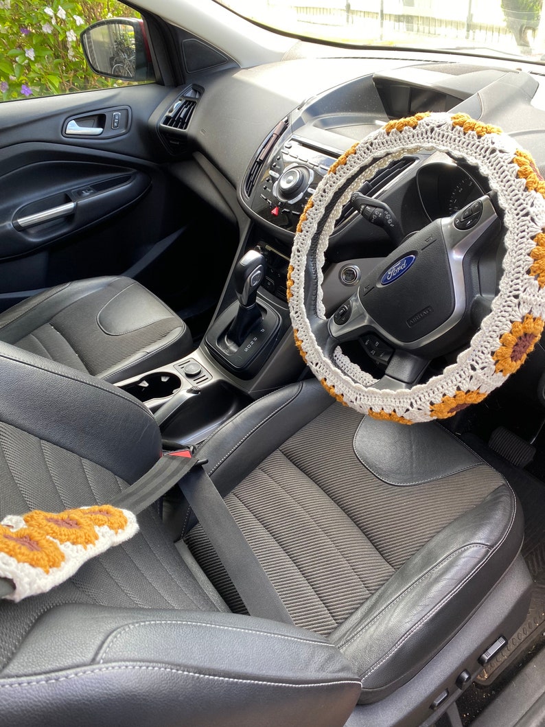 May include: The interior of a black car with a crocheted steering wheel cover. The cover is white with brown accents. The car has black leather seats and a black dashboard.