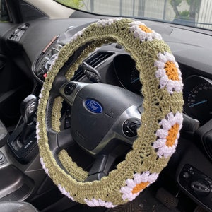 May include: A green crocheted steering wheel cover with white daisy flowers and orange centers. The cover is on a black steering wheel with the word "Ford" in the center.