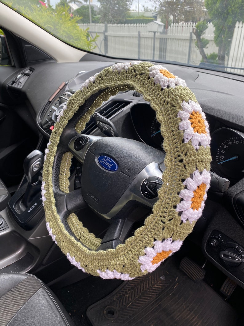 May include: A green crocheted steering wheel cover with white daisy flowers and orange centers. The cover is on a black steering wheel with the word "Ford" in the center.