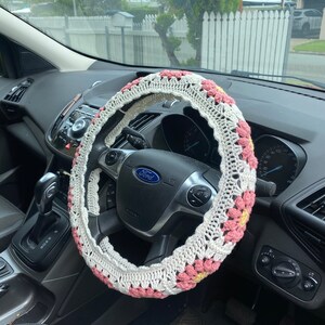 May include: A white and pink crocheted steering wheel cover with a floral design. The cover is on a black steering wheel with a Ford logo.
