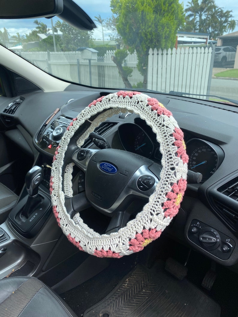 May include: A white and pink crocheted steering wheel cover with a floral design. The cover is on a black steering wheel with a Ford logo.