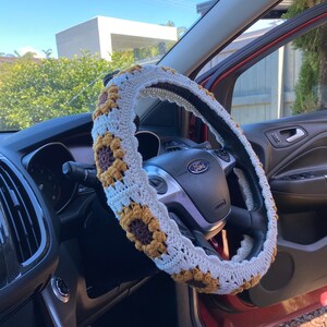 May include: A white crocheted steering wheel cover with brown and yellow sunflower accents. The cover is on a black steering wheel with the word "Ford" in silver lettering.