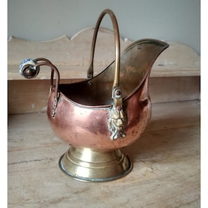 Peut inclure: Un seau à charbon en cuivre et en laiton avec une poignée et une tête de lion décorative sur le côté. Le seau est posé sur une surface en bois.