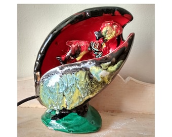 Può includere: Una lampada in ceramica a forma di conchiglia con un interno rosso e tre pesci all'interno. L'esterno della conchiglia è nero, verde e giallo.