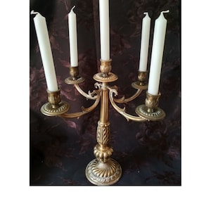 May include: A gold-toned metal candelabra with five arms and five white candles. The candelabra has a decorative base and ornate details.