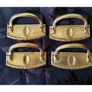 May include: Four antique brass drawer pulls with ornate designs. Each pull has a rectangular plate with a raised oval in the centre and a decorative border. The pulls have a curved handle.