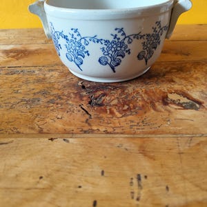 Op de afbeelding: Witte keramische kom met een blauw bloemenpatroon en twee handvatten. De kom staat op een houten oppervlak.
