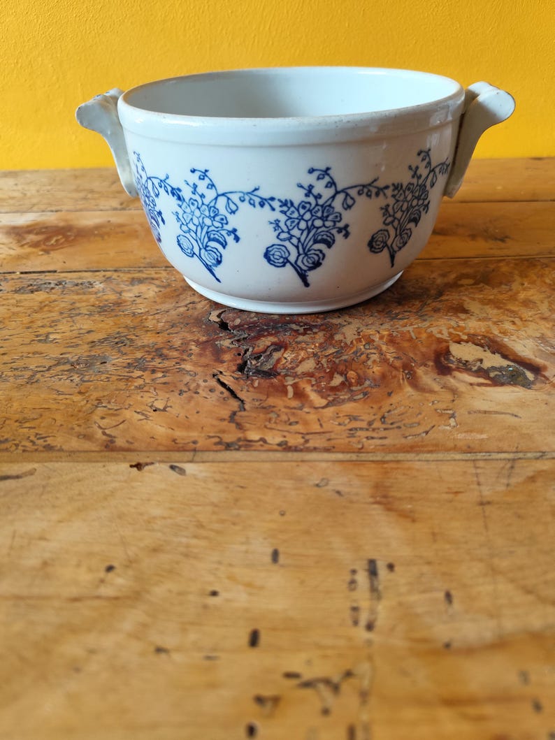 Op de afbeelding: Witte keramische kom met een blauw bloemenpatroon en twee handvatten. De kom staat op een houten oppervlak.