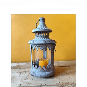 Puede incluir: Un farol de metal gris desgastado con un acabado envejecido, que presenta un borde decorativo con patrón de hojas a lo largo de la parte superior e inferior de los paneles de vidrio. Una pequeña vela de pilar amarilla se encuentra dentro.