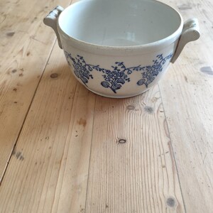 Op de afbeelding: Een witte keramische kom met blauwe bloemenmotieven en twee handvatten. De kom staat op een houten oppervlak.