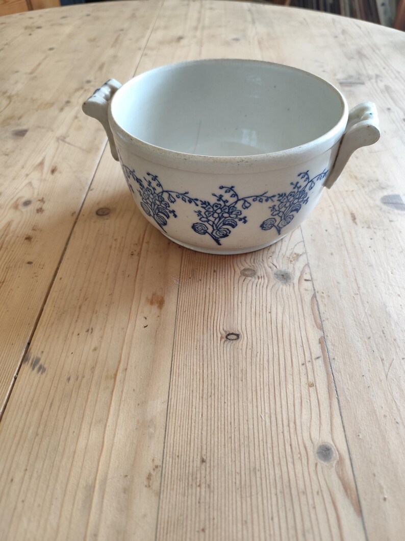 Op de afbeelding: Een witte keramische kom met blauwe bloemenmotieven en twee handvatten. De kom staat op een houten oppervlak.