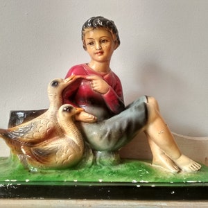 Puede incluir: Una figurita de un niño pequeño sentado con dos gansos. El niño lleva una camisa roja y pantalones azules. Los gansos son blancos con marcas marrones. La figurita está hecha de yeso y está pintada en colores realistas.