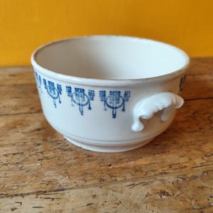 K&ouml;nnte beinhalten: Eine wei&szlig;e Keramiksch&uuml;ssel mit einem blauen und wei&szlig;en Blumenmuster am Rand. Die Sch&uuml;ssel hat zwei kleine Griffe an den Seiten.
